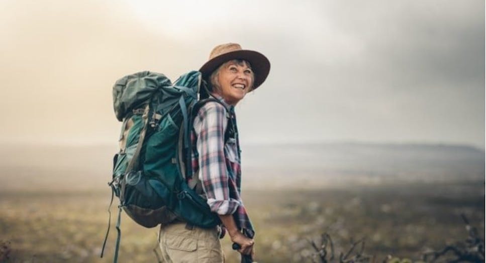 A more senior lady hiking with a green backpack and brown hat. She is smiling.