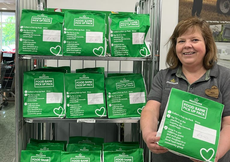 A female Morrisons staff member holds a green 'food bank pick up pack' while standing by a shelf full of similar bags.