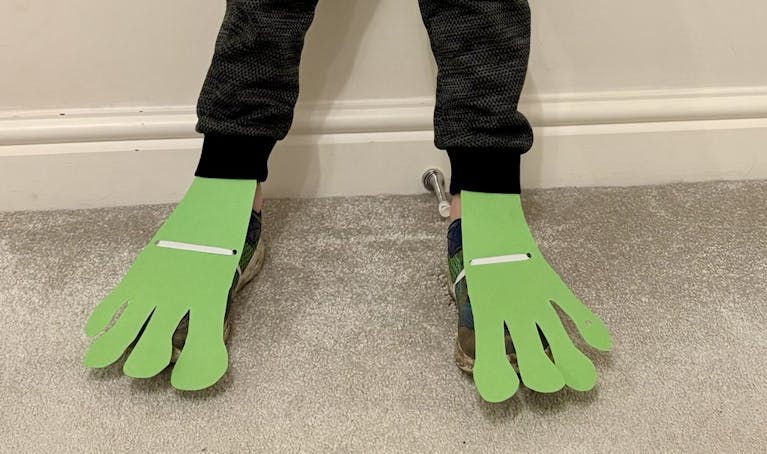 Image shows a child's feet with some home made 'frog feet' made out of green paper attached with elastic in the same way a ballet shoe might be attached to a foot
