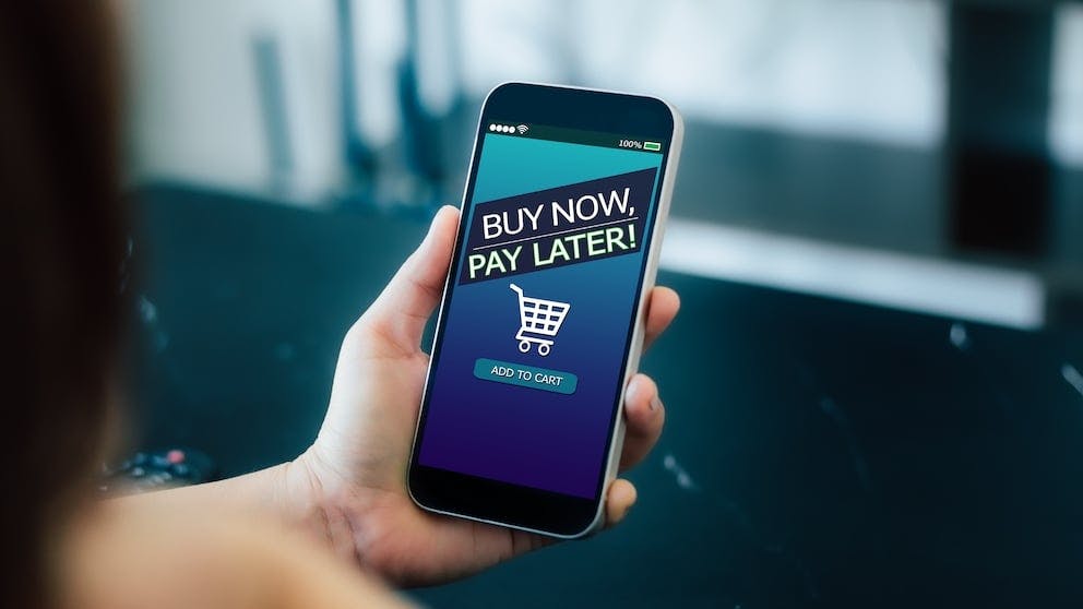 Mobile phone being held. On the screen it displays a message saying: 'Buy Now, Pay Later' and featuring a shopping trolley.