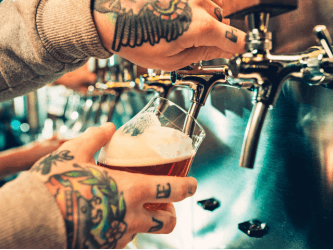 Close up of a glass of beer being poured by someone with tattoos on their hands.