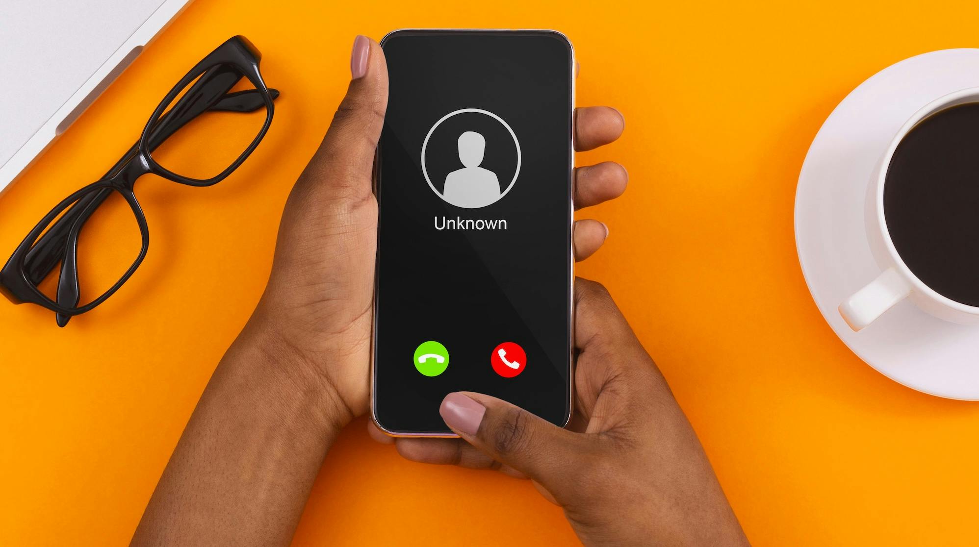 An orange desk provides the backdrop to two hands holding a mobile phone, with a call from an unknown caller coming through.
