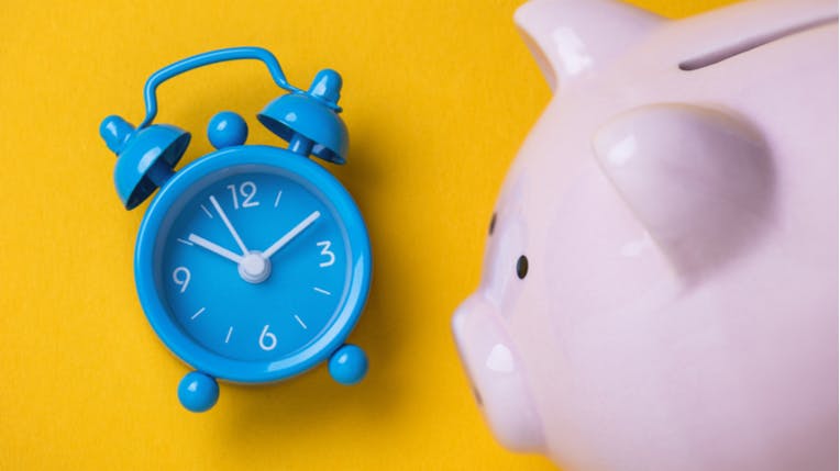 Photo of a blue alarm clock next to a pink piggy bank on a yellow background.