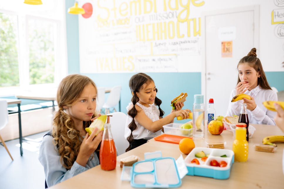 Image shows three young children - all girls - in a brightly lit classroom eating breakfast and lunch items including fruit a sandwich.