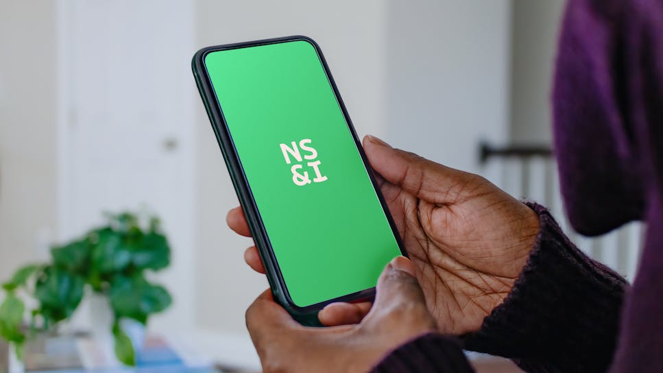 A person holding a phone with the NS&I logo showing on the screen.