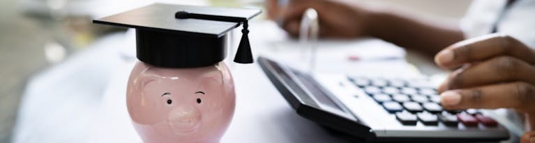 Stock image showing a piggy bank wearing a graduation cap while in the background a man types on a calculator