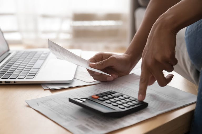 Someone's hands can be seen using a calculator which sits on some paperwork. A laptop stands open nearby.
