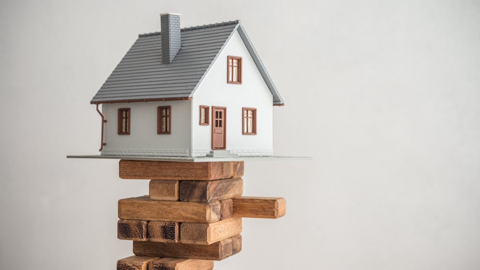 Scale model of a house balancing precariously on a wooden block tower