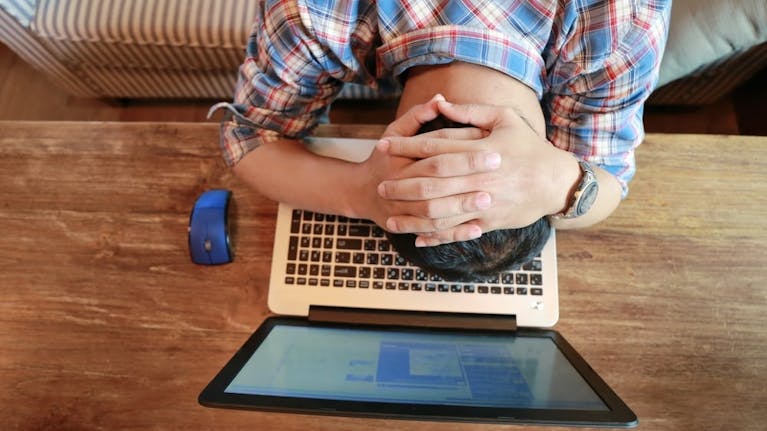 Man sitting on sofa with a laptop and mouse on the table in front of him, with his head on the keyboard, facing down, and his hands clasped behind his head.
