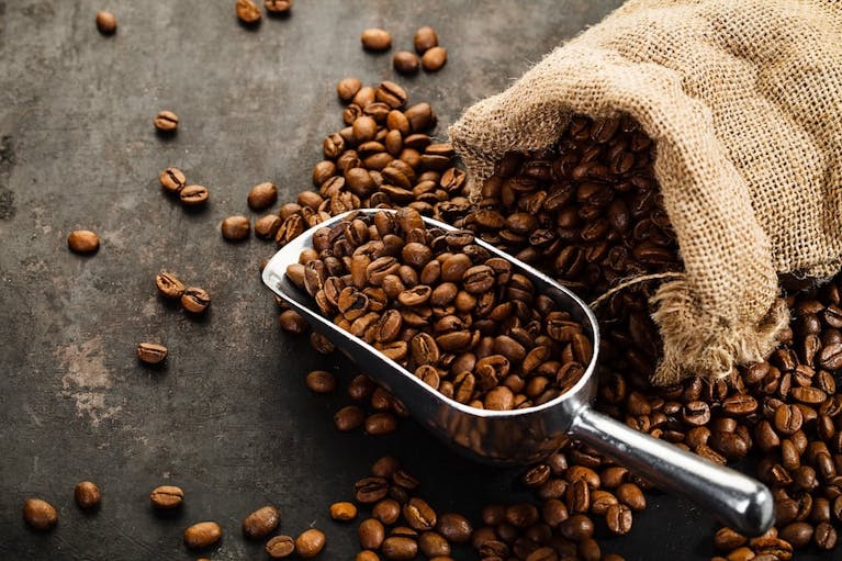 A hessian sack spilling coffee beans onto a black surface. A silver scoop sits next to it