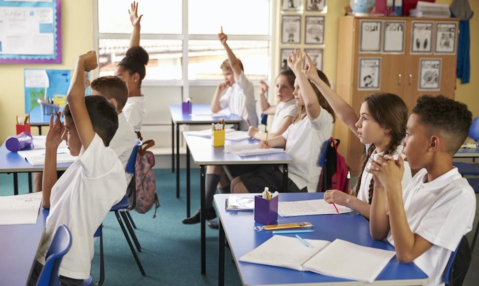 Children in a classroom sitting at blue desks, some have their hands up asking to answer a question.