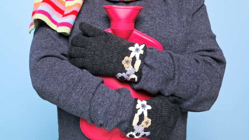 Close up of a person wearing a jumper, scarf and gloves hugging a hot water bottle.