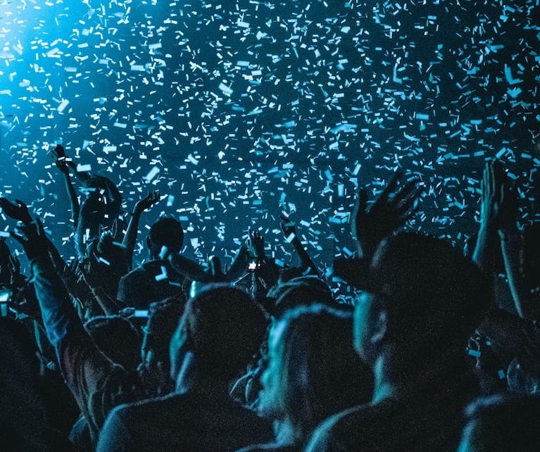 An audience watches a concert in a dark venue as white and blue streamers fall from above
