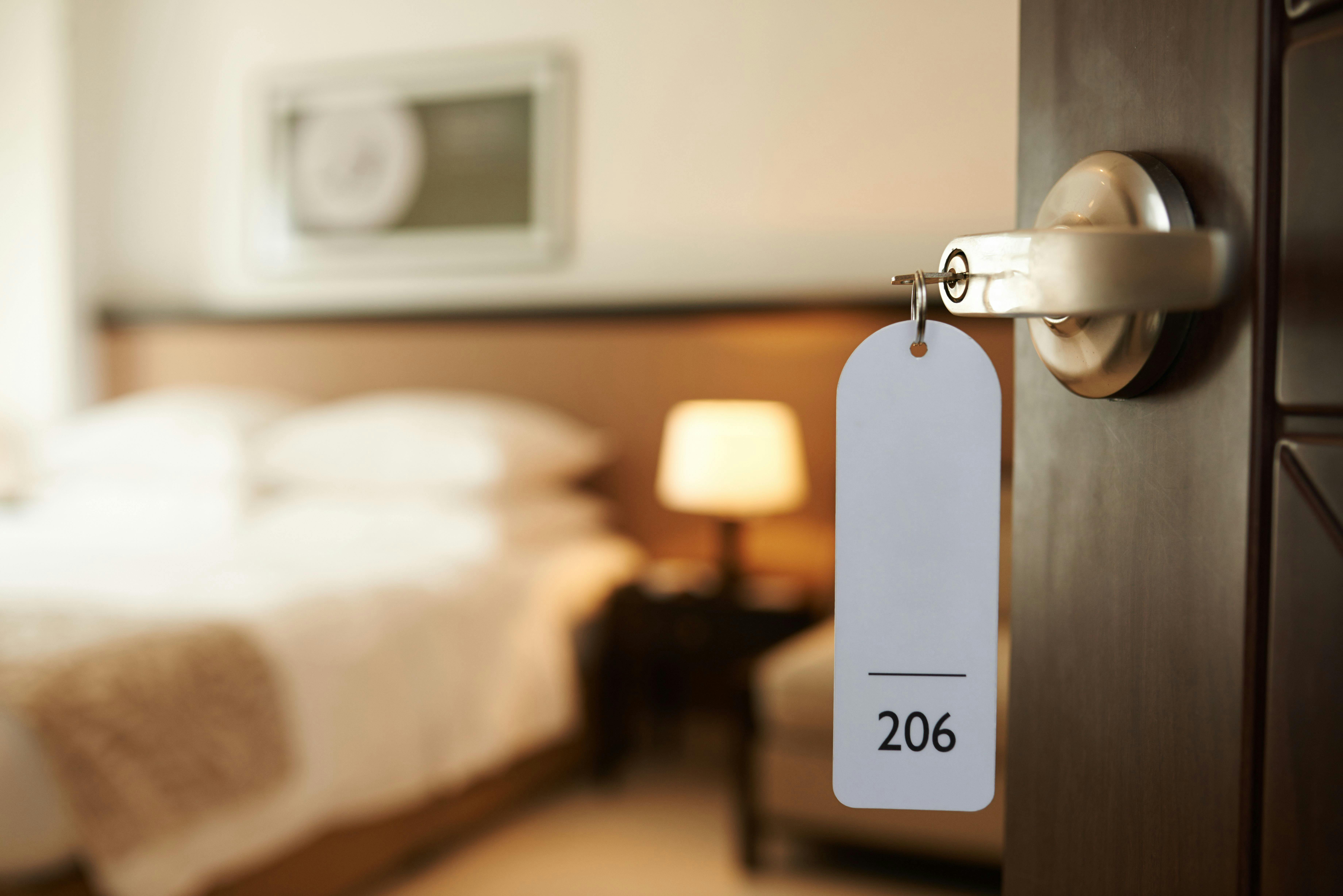 A blurred hotel room through an open door, the focus is on the key in the lock with the number '206' on the tag hanging from it.